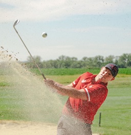 A Male USD Golfer hitting the ball out of the sand bunker.