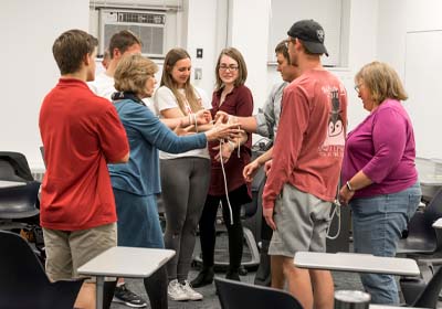 Students standing in a circle in a classroom, most are reaching their hands in for what looks like a hand-stack