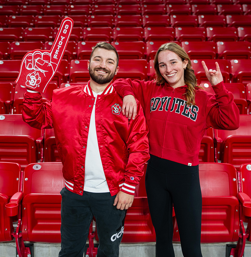 Two USD students wearing red coyote gear. One has her coyote hand in the air, while the other is sporting a go coyotes foam finger.