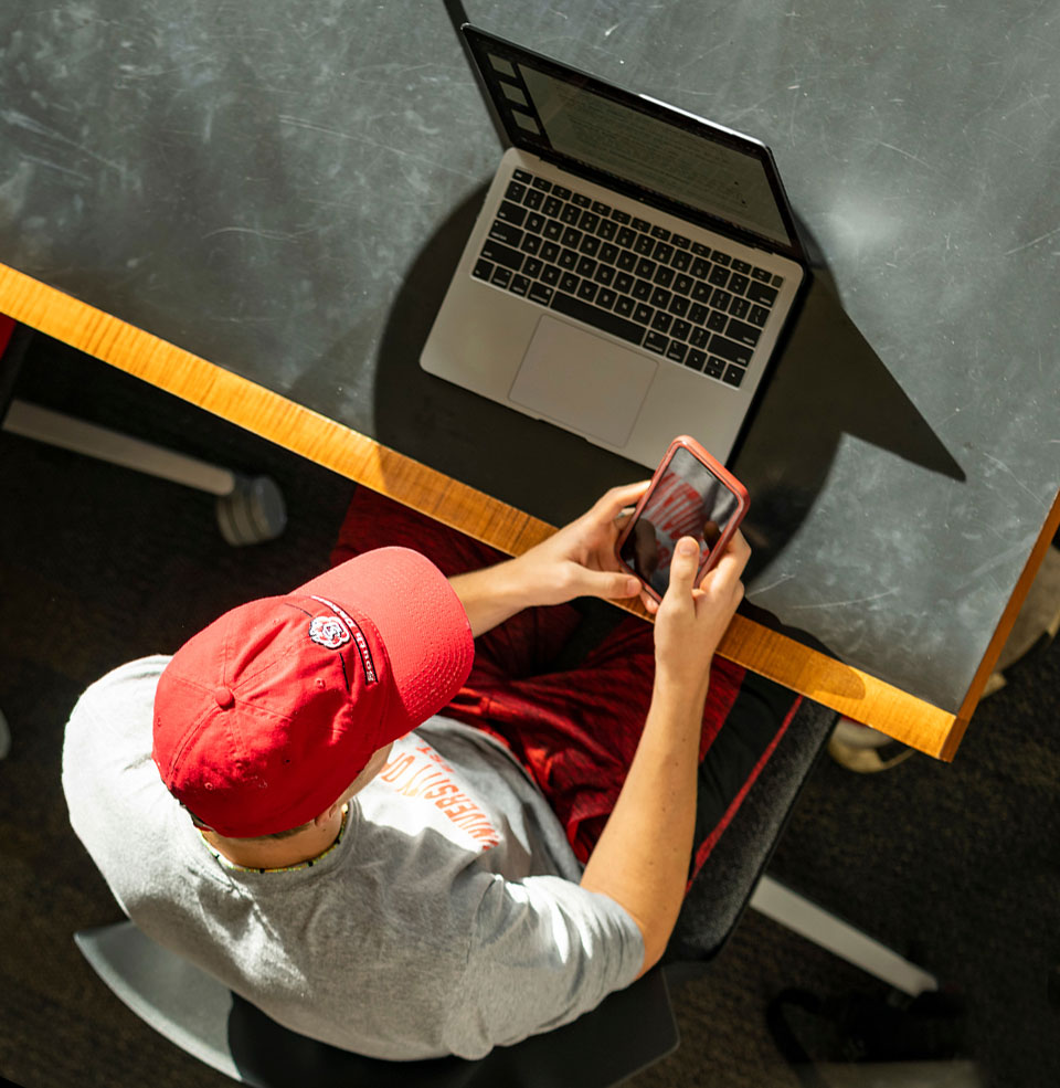 Student sitting at a laptop on a desk looking at a phone