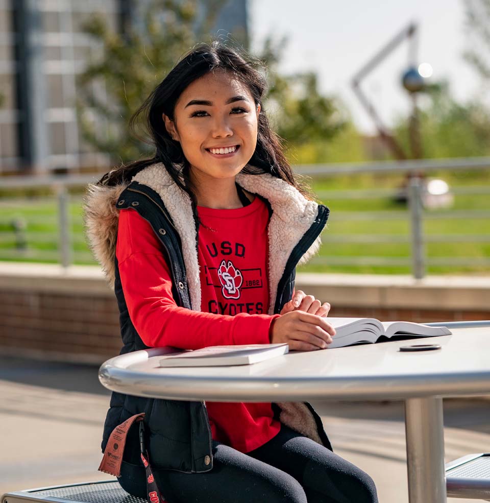 Female Student Sitting Outside at Table Smiling.