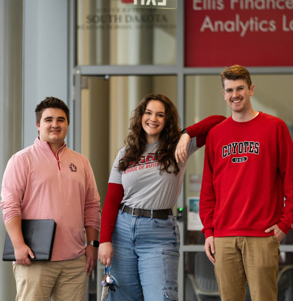 Three Business students smiling in front of the Ellis Finance Lab.