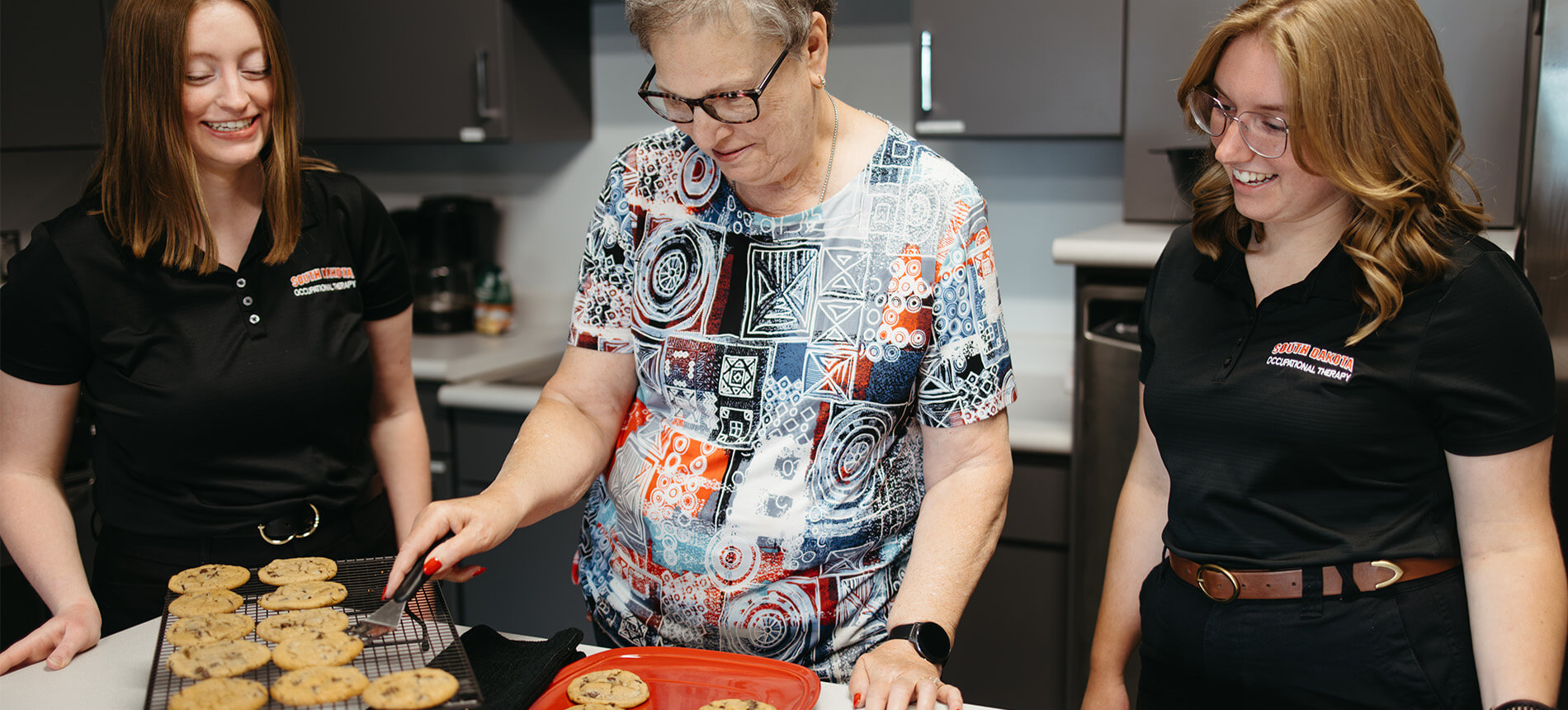two OT students helping a patient with fine motor skills by transferring cookies from a pan to a cooling rack