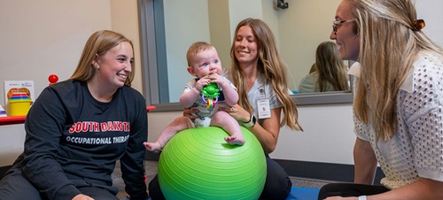 PT and OT students helping a baby sitting on a medicine ball in a childcare service location.