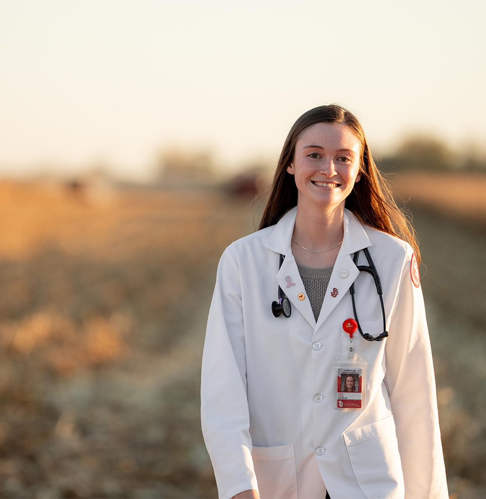 Student smiling in white coat and stethoscope, standing outside at dusk with a rural South Dakota background. 