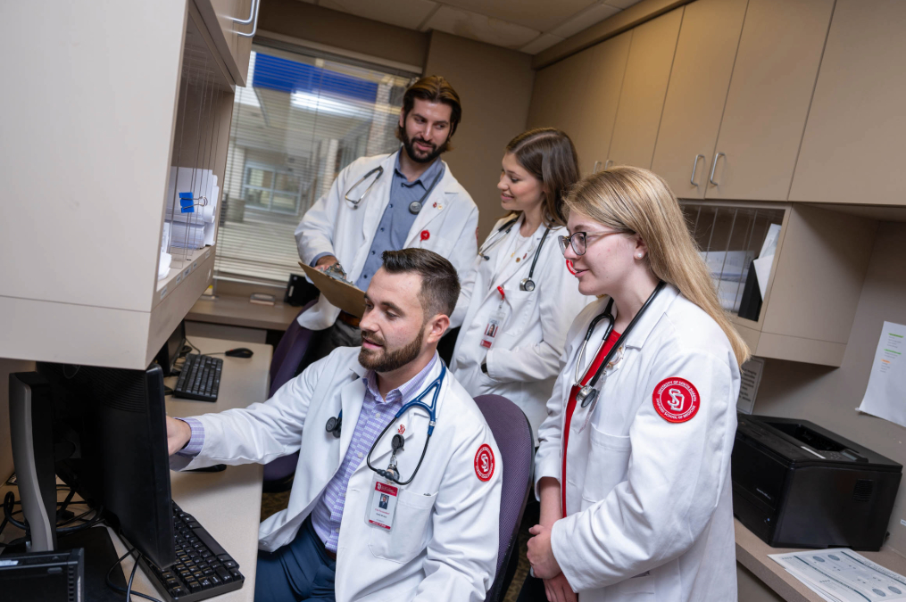 Four med students in white coats standing around a computer reading what's on screen.