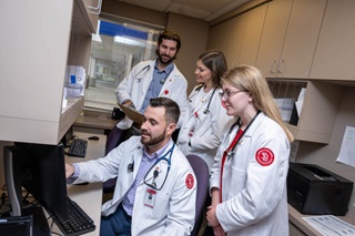 Four med students in white coats standing around a computer reading what's on screen.