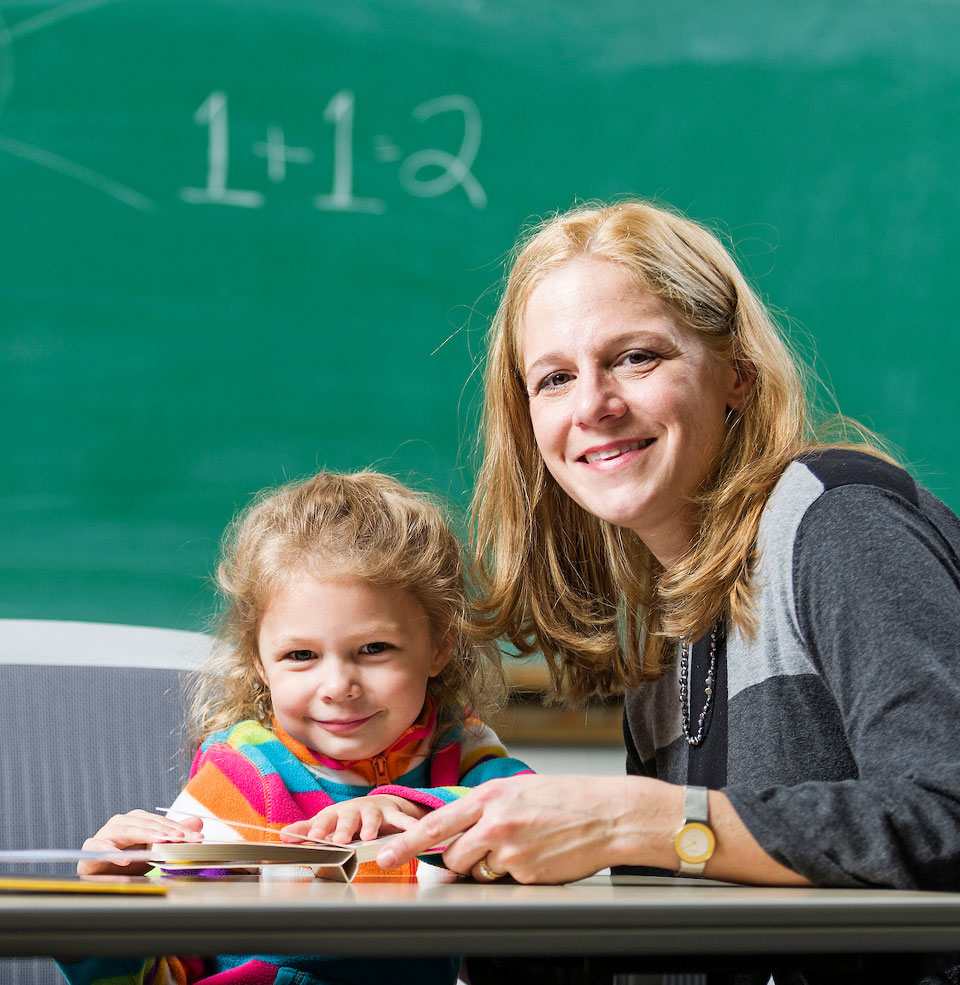 A teacher and younger student in a classroom near a chalkboard smiling at camera 