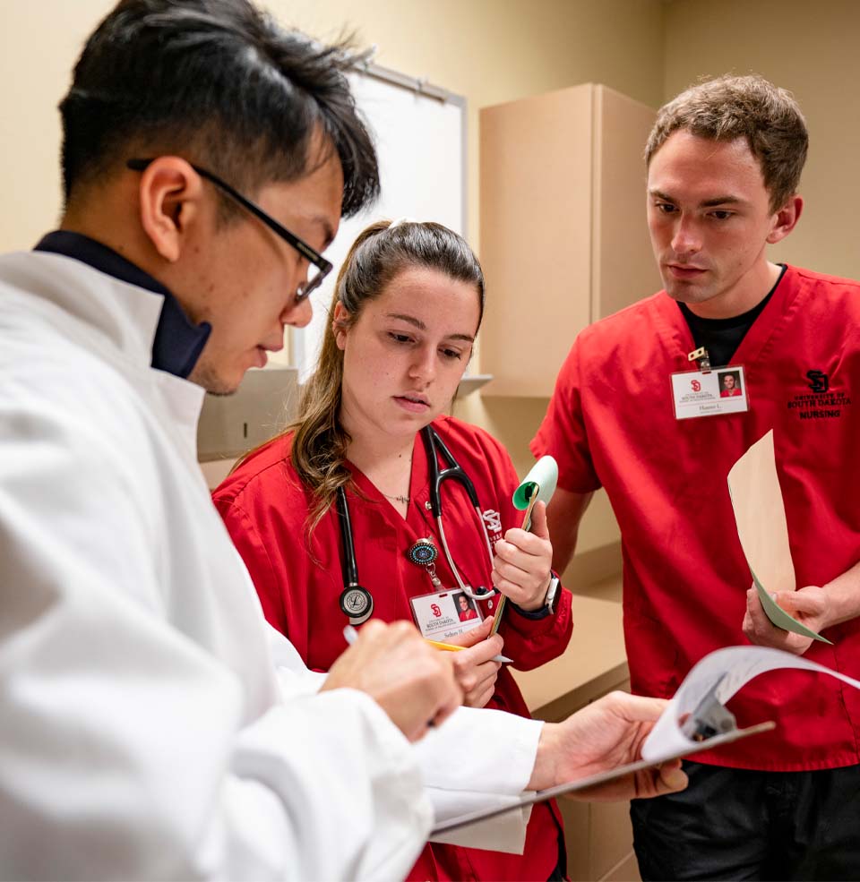 Two nursing students in red scrubs look at a clipboard with a medical student in a white lab coat.
