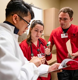 Two nursing students in red scrubs look at a clipboard with a medical student in a white lab coat.
