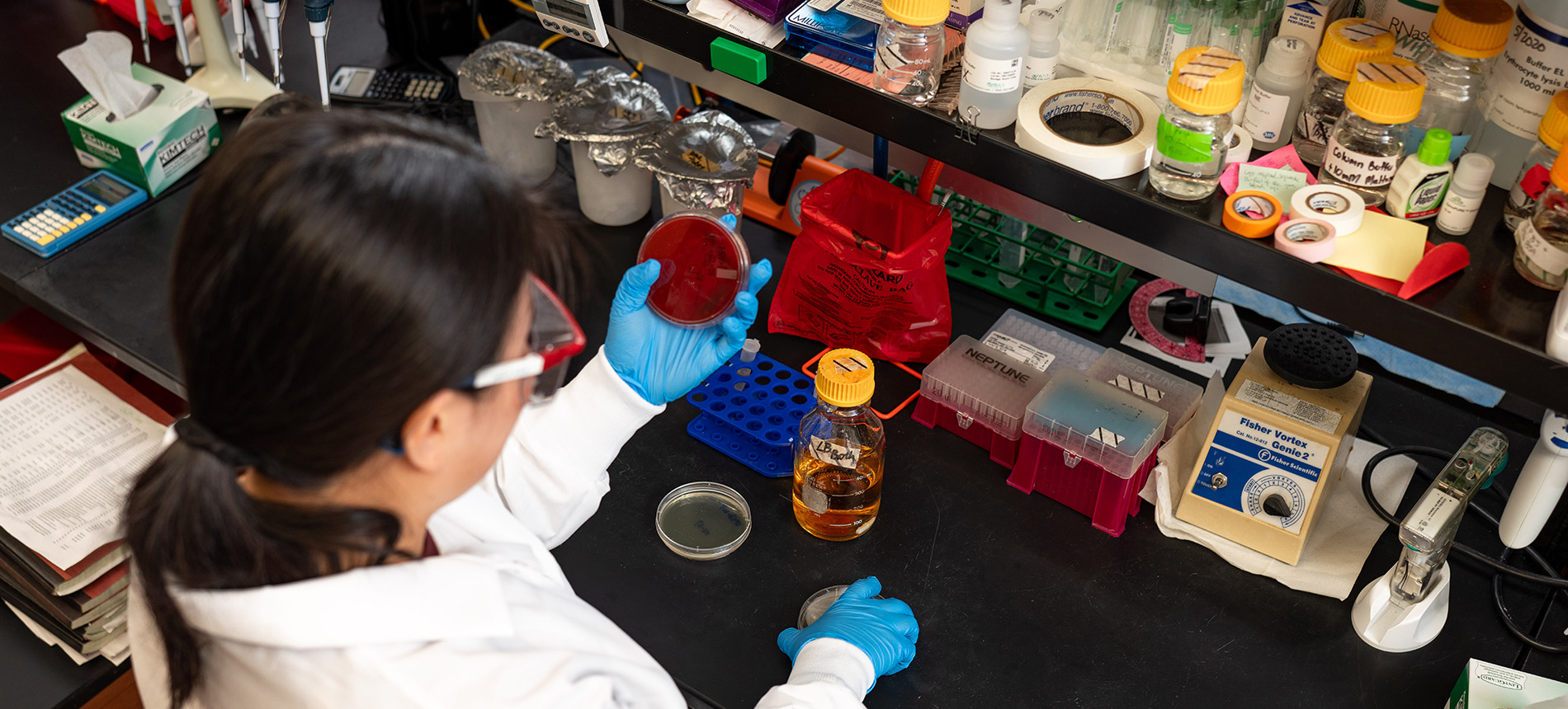 Person in lab coat and goggles with an array of chemicals and lab equipment on a table in front of them. 