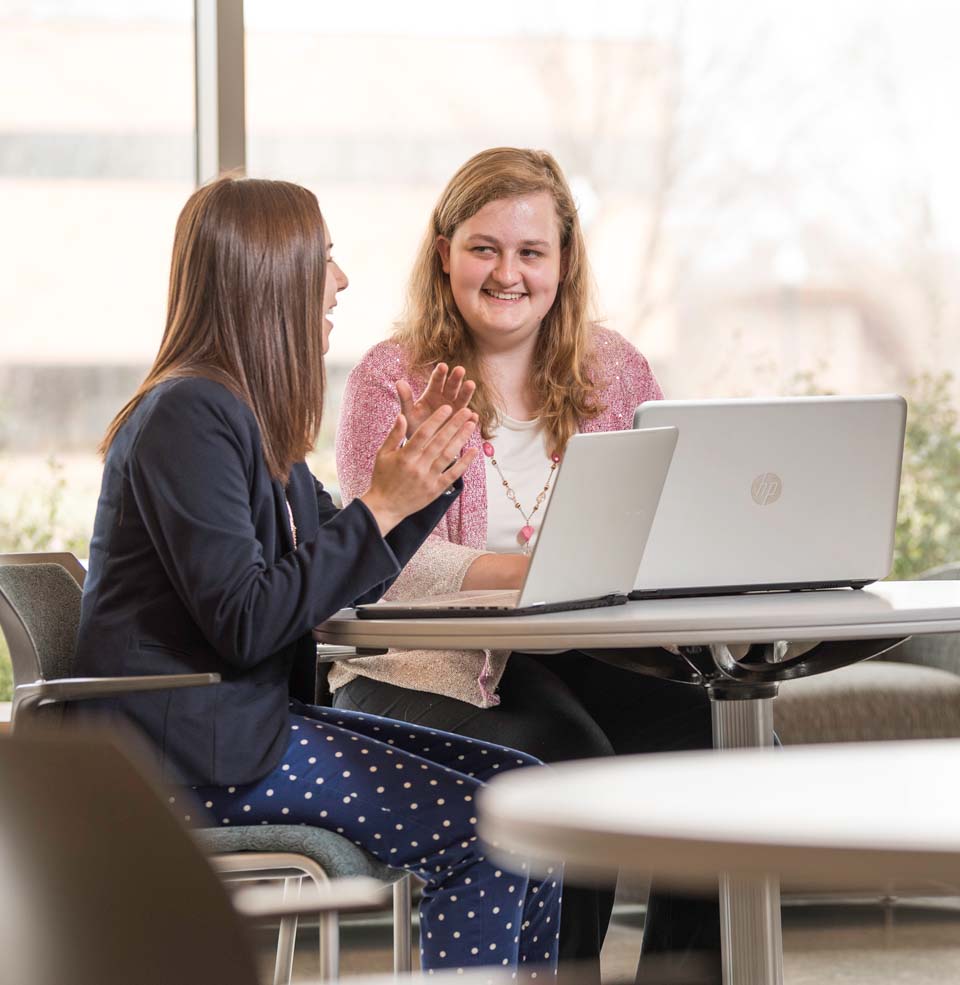 Two Female Students Interacting at a Table with Their Laptops.