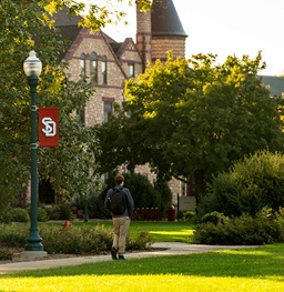 Student walking on outside on campus next to lamp and green trees.