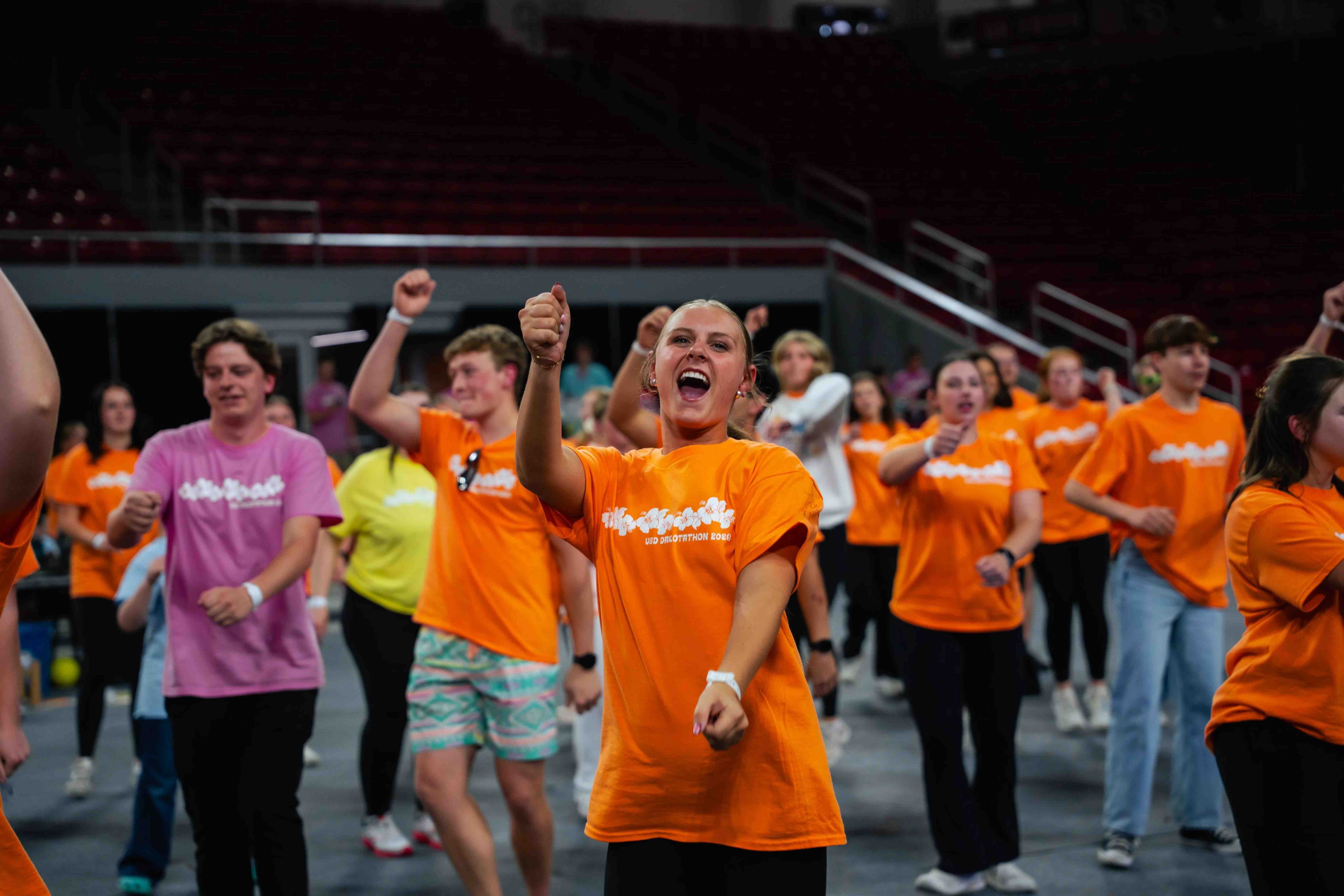 A young woman in an orange T-shirt leads a group of people dancing in a large arena.