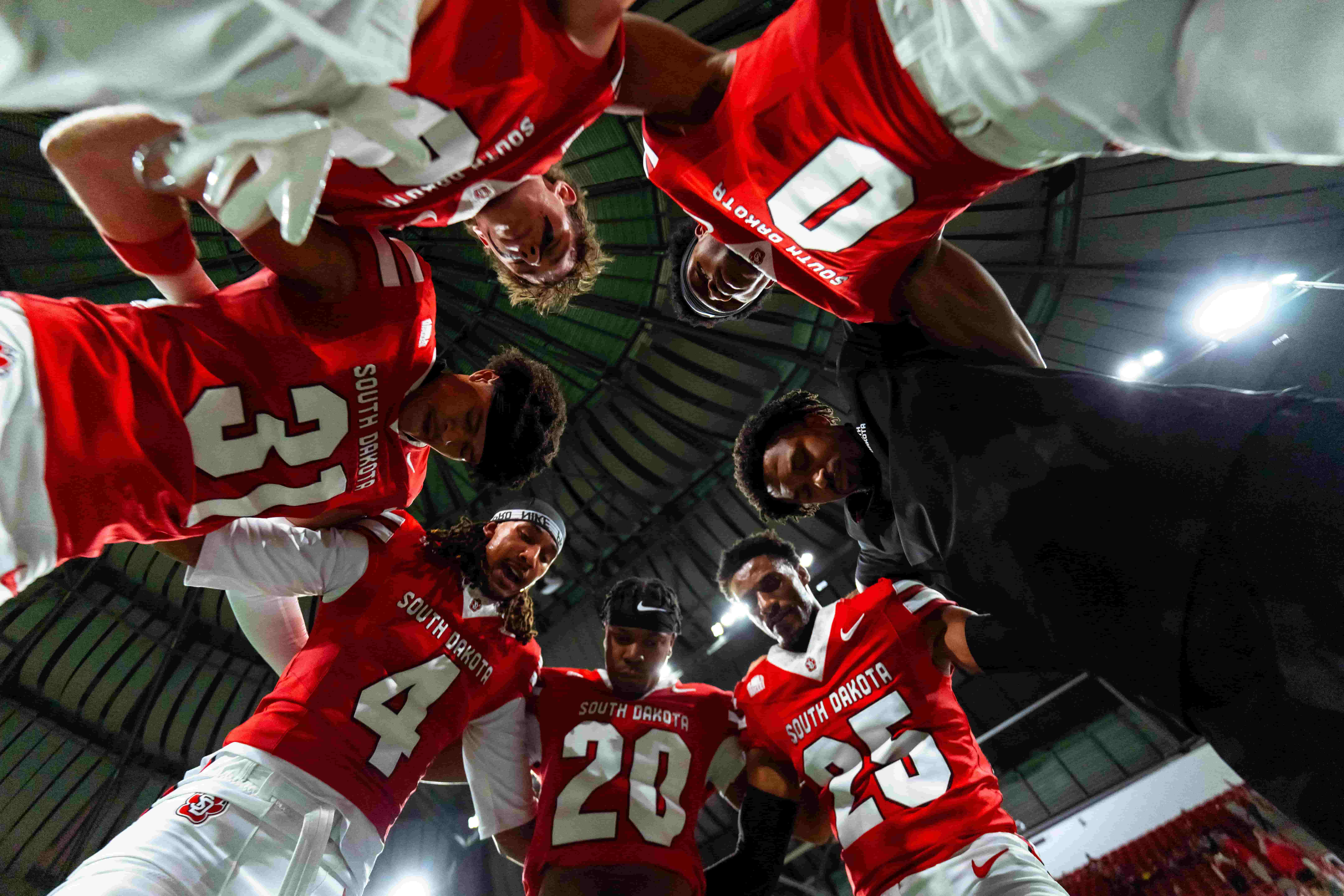 South Dakota football players in red jerseys huddle beneath stadium lights, looking focused and united before a game.