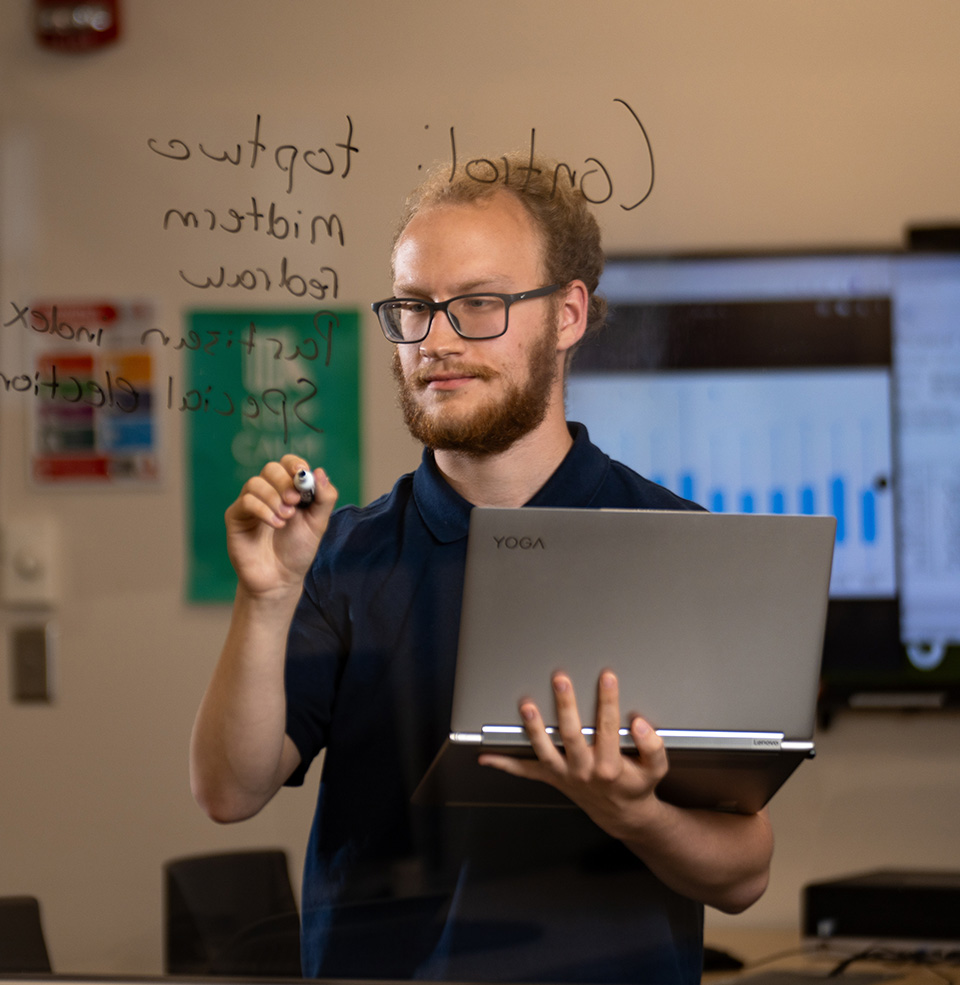 A student writing on glass with a dry erase marker working on their research project with a bar graph in the background