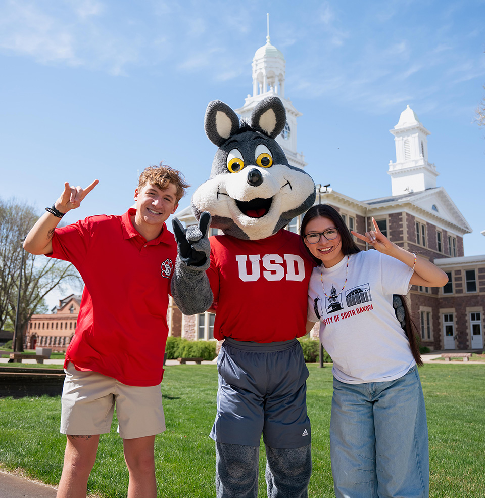 Charlie Coyote with students holding up a yote hand outside of Old Main on USD Vermillions campus