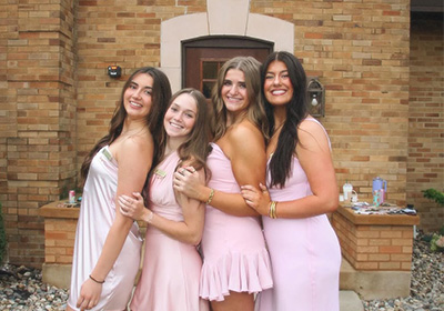 Four sorority sisters dressed in pink stand hugging eachother in front of their house. 