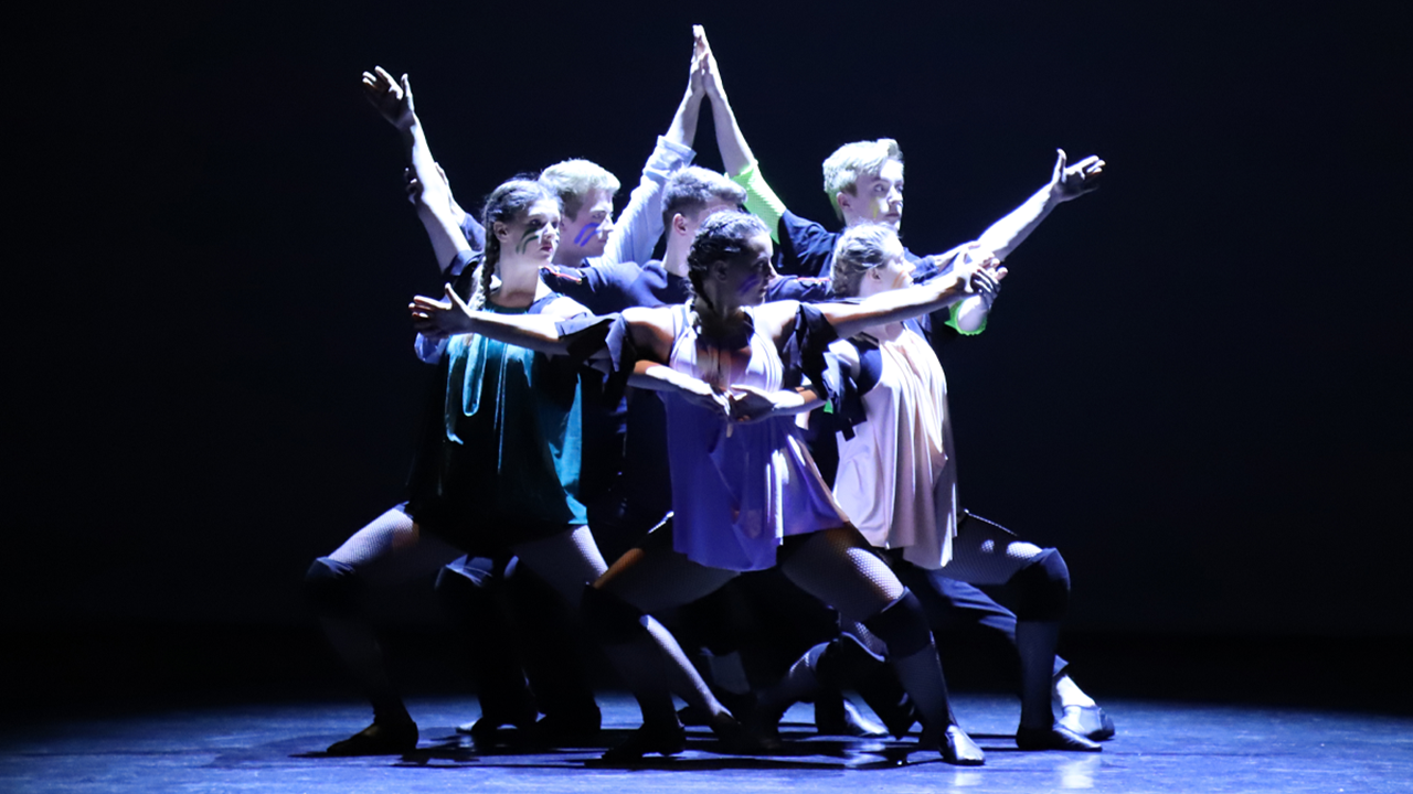 Dancing students crouch and stand together with their arms locked in an artistic and interesting way. The lighting is dark with a hint of blue and purple light. They all look to their left.