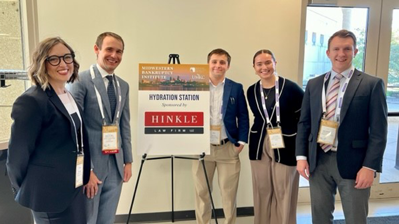 Four students and a professor stand next to a posterboard at a conference and smile for a photo. They are dressed in professional wear.