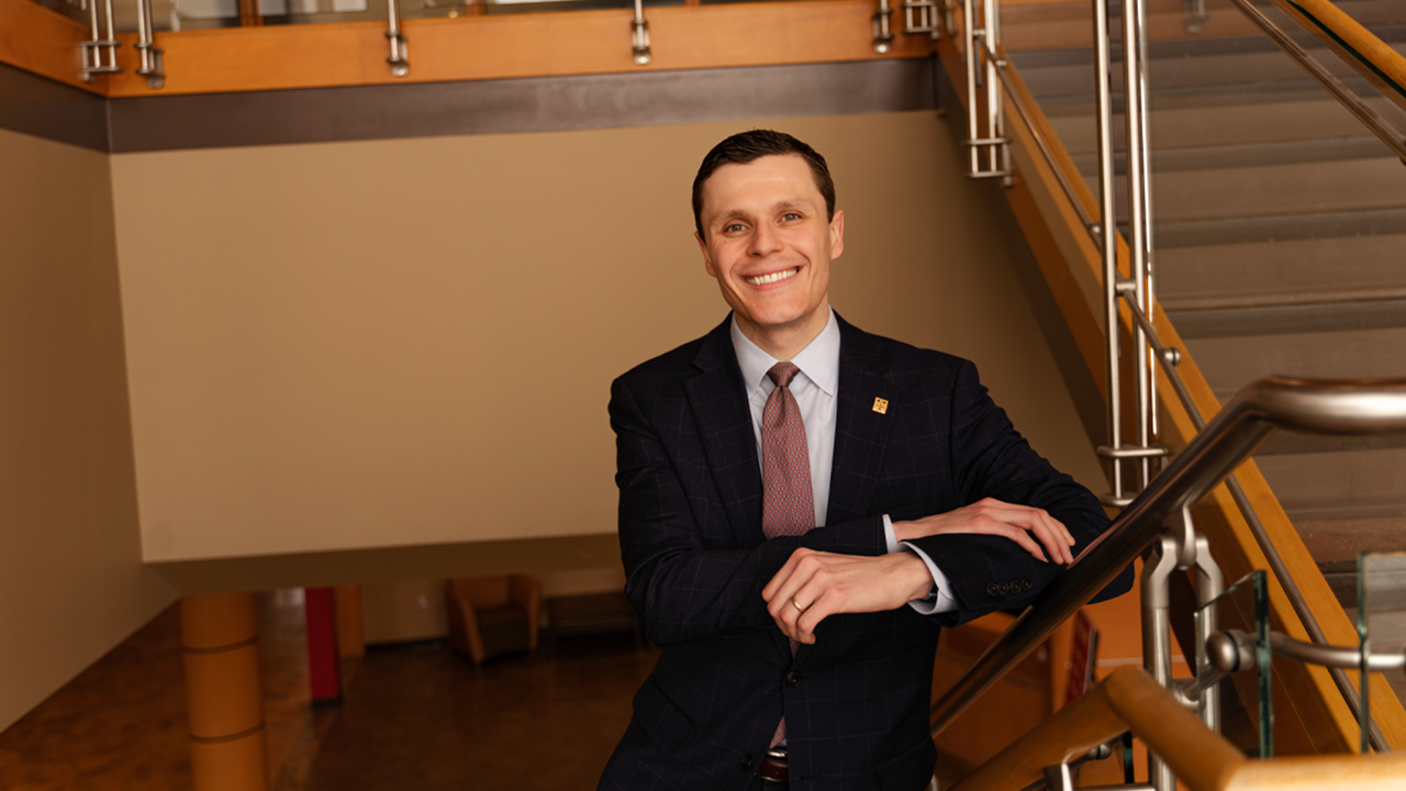 Dzenan Berberovic stands on a flight of stairs with his arms crossed. He is smiling and wearing a suit and tie.