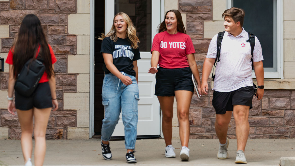 Three students walk out the doors of Old Main, waving and smiling to students who are walking in. They are all wearing Charlie's gear.