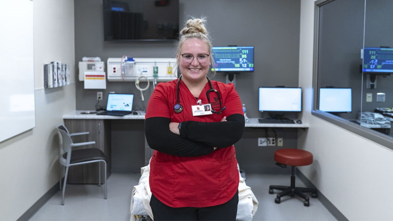 A USD student in scrubs, standing in front of computers. A USD student in scrubs, standing in front of computers.