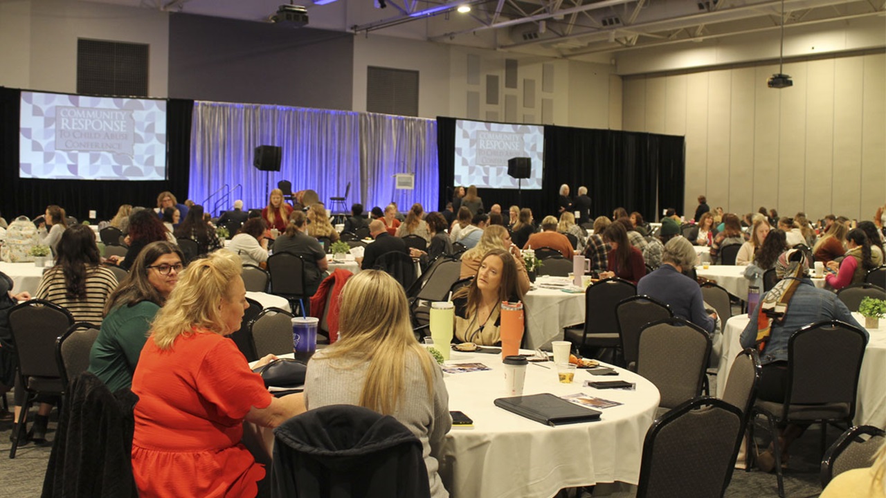 CPCM - Child Abuse Prevention Conference 2023 Dozens of professionals seated at round tables in a convention hall setting wait for the next speaker to ascend to a well-lit stage flanked by large display screens.