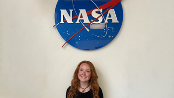 Maddie Rozmajzl stands against a beige wall for a photo. A sign with NASA's logo on it hangs above her.
