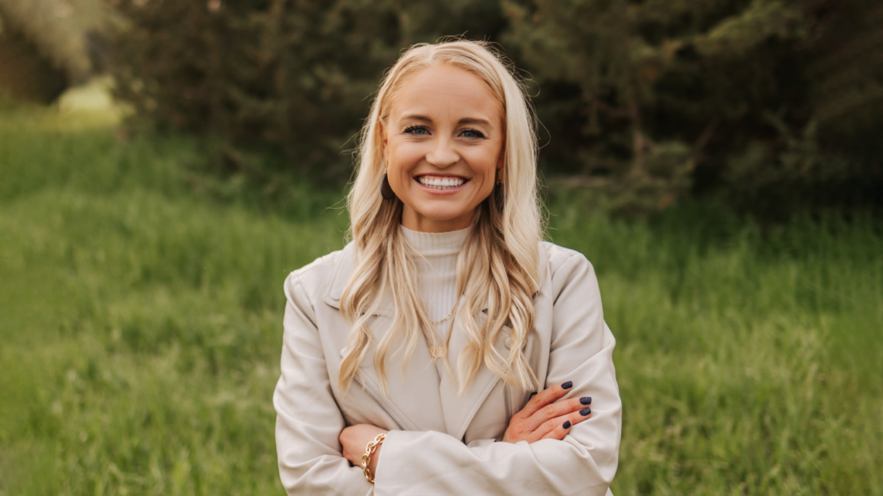 Rachelle Schmidt stands and smiles with her arms crossed. She is standing in a field of green grass.