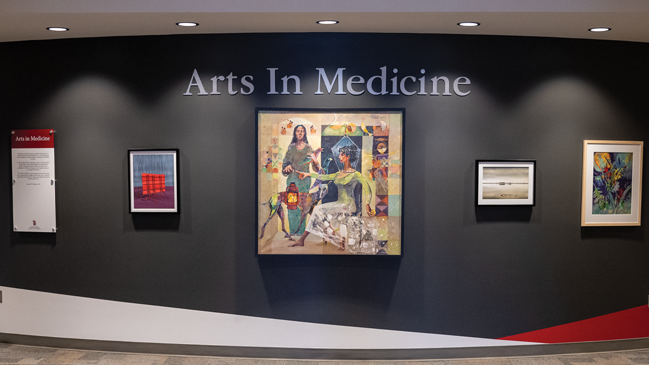 A curved wall that has five framed artwork pieces hanging on it. Above the artwork is the words Arts In Medicine.