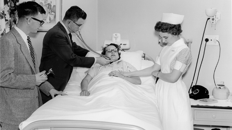 A black and white photo of medical professionals in the 1950s helping a patient in bed.