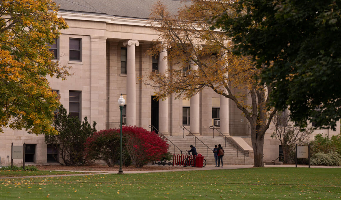 The College of Arts & Sciences sits on the USD campus, surrounded by fall trees and green grass, along with a couple of students.