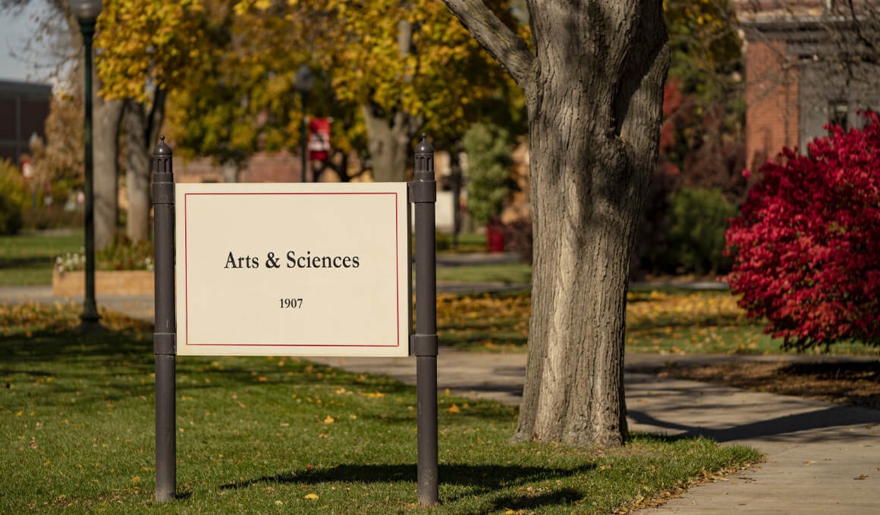 A sign that says "Arts & Sciences" sits in front of a tree on the USD campus, with fall bushes in the background.