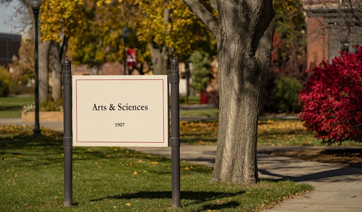 A sign that says "Arts & Sciences" sits in front of a tree on the USD campus, with fall bushes in the background.