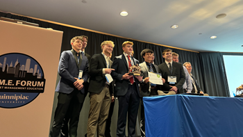 USD students stand behind a table, wearing professional attire, with a banner for the GAME Forum, celebrating with a trophy and a certificate.
