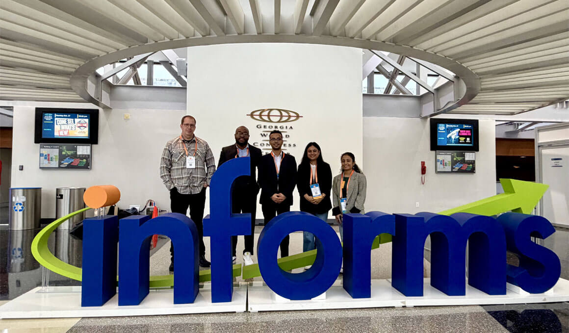 M.S. in Business Analytics students, and their mentor, stand behind a blue and green "INFORMS" sign at the INFORMS Annual Meeting.