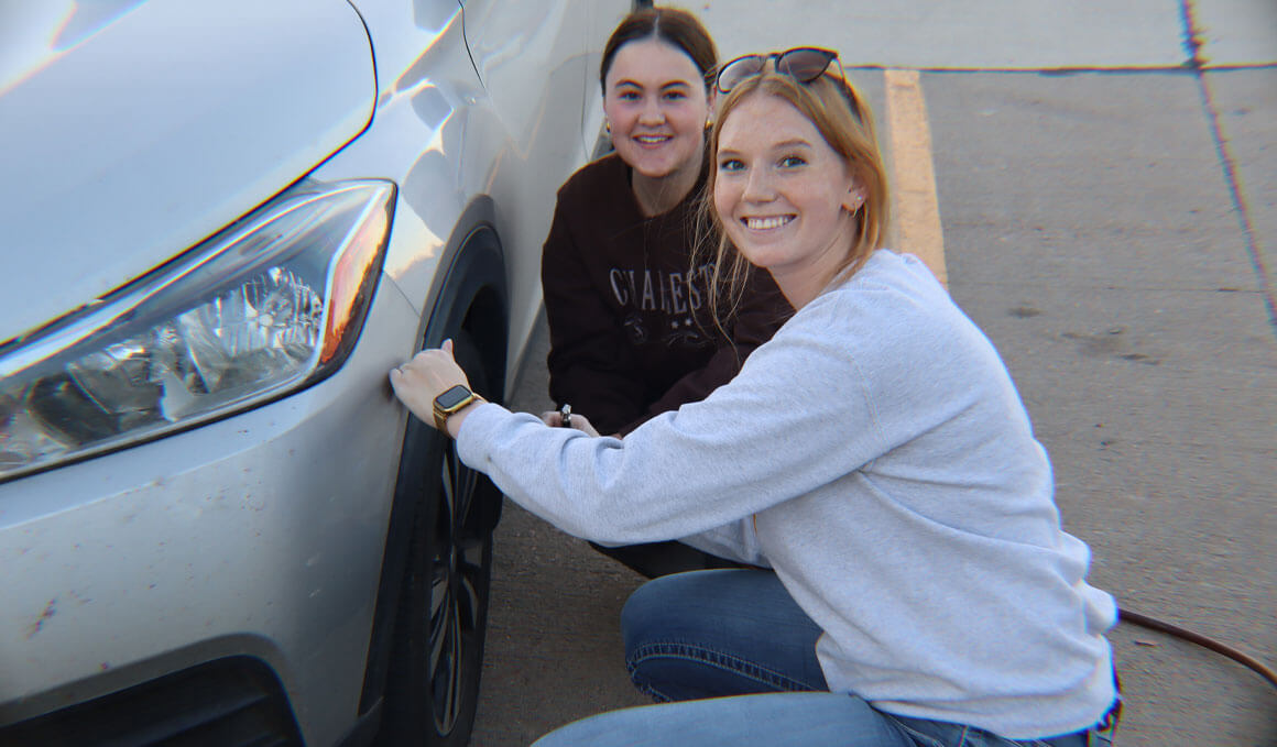 Two students sit on the driver's side of a white car, holding the hose to an air pump, and smiling at the camera.