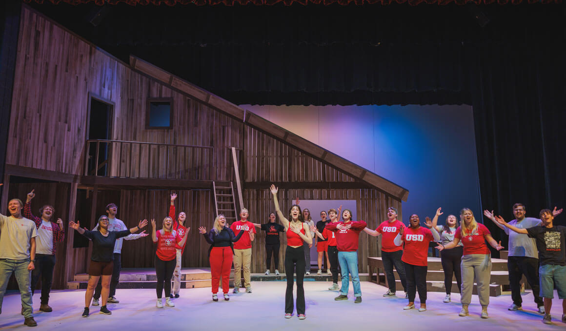 Members of USD's theatre stand on stage, with their hands pointing up; a wooden building is in the background.