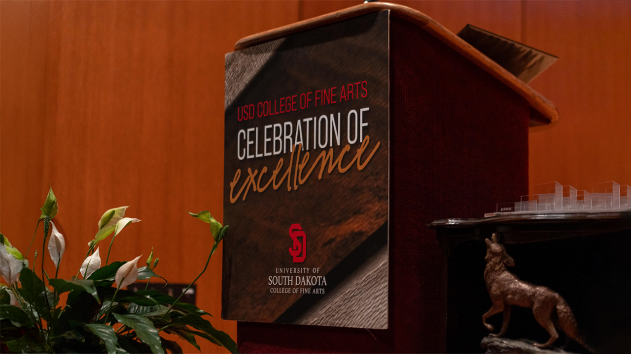 A black podium with the words "USD College of Fine Arts Celebration of Excellence" and the USD logo sit on a stage, next to a green plant and a table holding a miniature Coyote legacy statue.