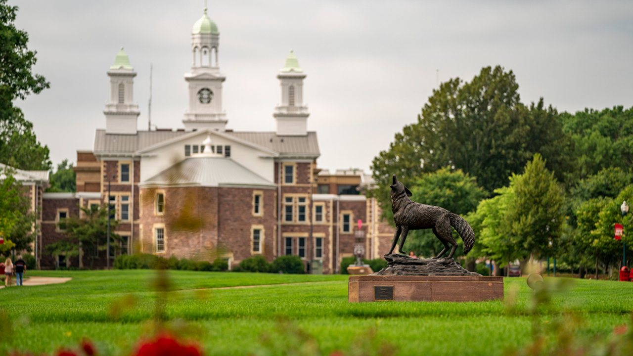 A bronze statue of a howling coyote stands in the foreground of a grassy lawn with a large, multi-story brick building in the background under a cloudy sky.