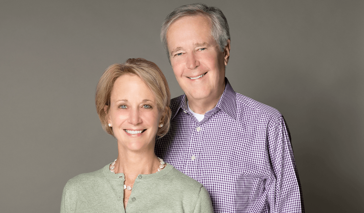 James and Deborah Fallows stand together, facing the camera with wide smiles, against a gray backdrop.