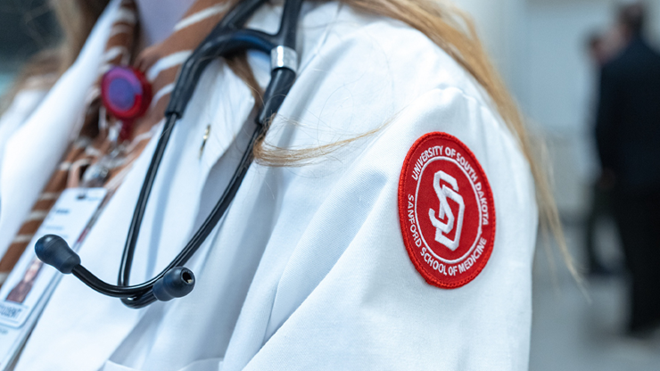 A USD Sanford School of Medicine student wears a white coat with the SSOM badge, in red, on the white coat's sleeve.