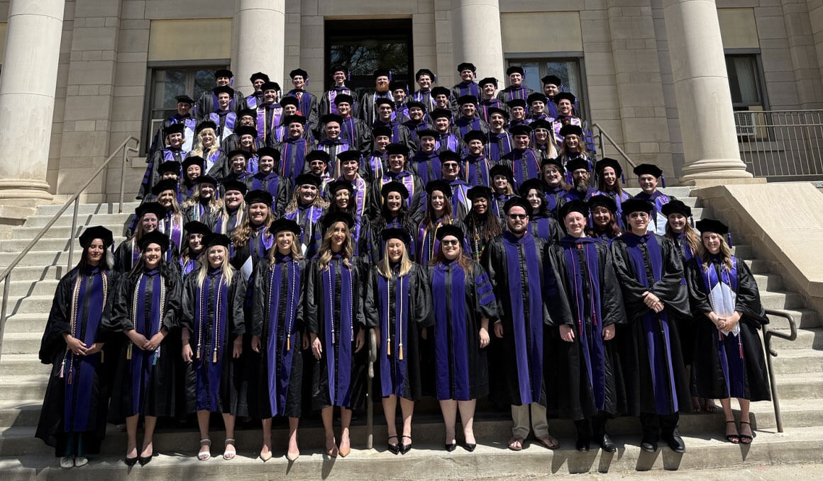 A large group of graduate students in black and purple academic robes pose on the steps of a building for a hooding ceremony photo.