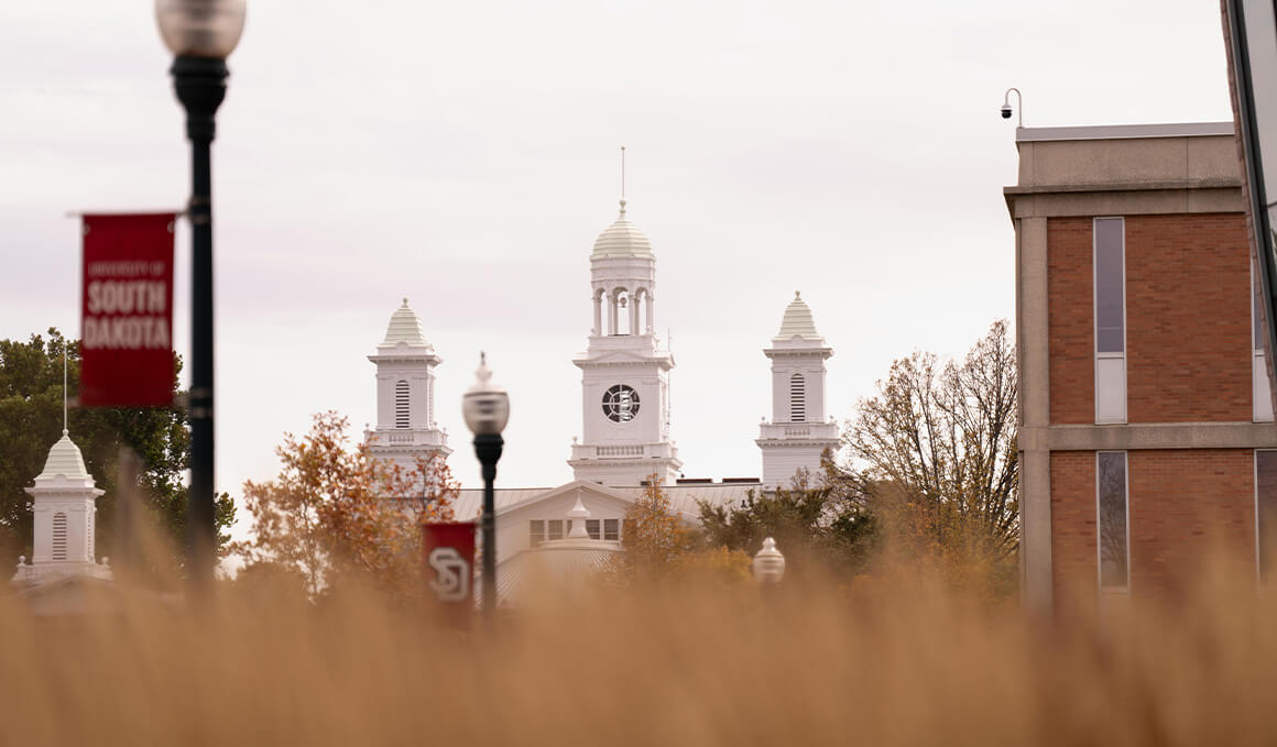 View of the USD campus featuring the white cupolas of Old Main framed by red USD banners, autumn trees and golden grasses in the foreground.