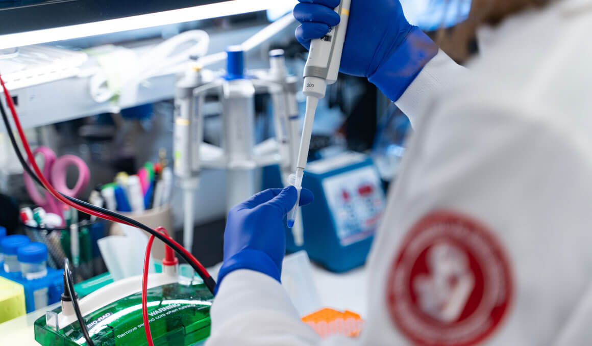 A USD Sanford School of Medicine student, wearing a white coat embroidered with the USD SSOM logo, works in a laborarory, holding test tubes.