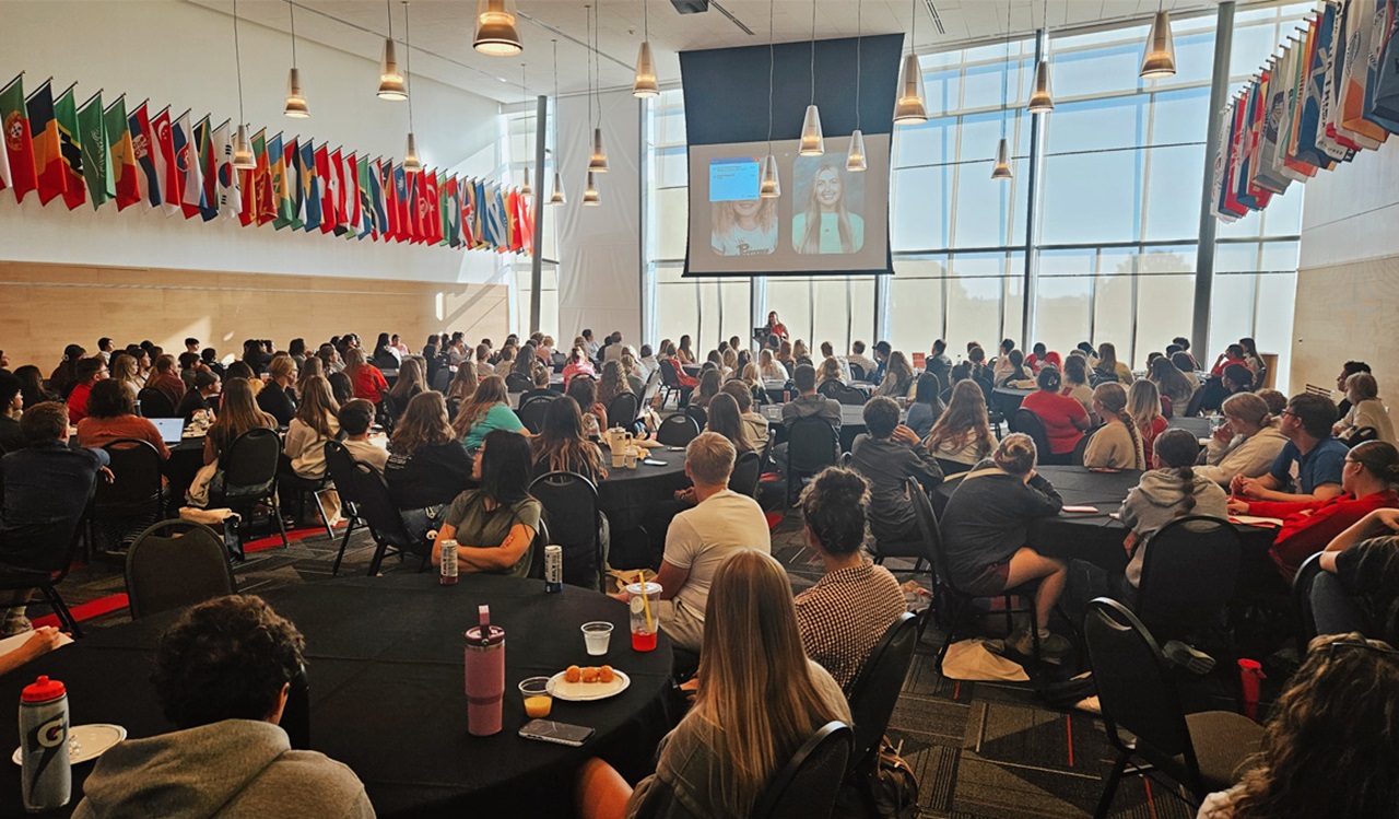 The USD School of Education hosts students and educators as they gather in a bright, flag-lined conference room to watch a presentation projected at the front.