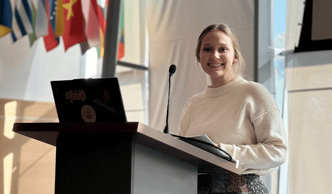 Jeryn Mediger-Lurz, a 2023 USD graduate and Teacher Pathway alum shares her journey at the Teacher Pathway Day, standing at a black podium, with a microphone and laptop in front of her, looking towards her left and smiling at the camera.