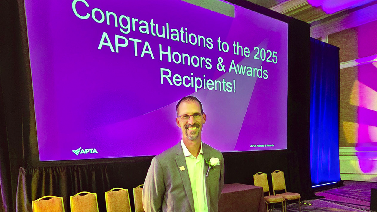 USD's Kory Zimney, DPT, Ph.D., stands in front of a large T.V. screen that has a purple background with white words that read "Congratualtions to the 2025 APTA Honors & Awards Recipients!"