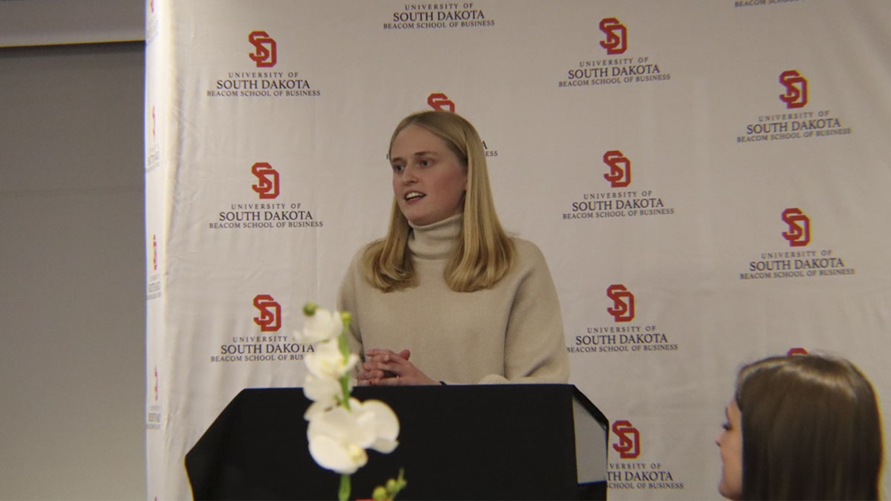 Elyse Whittemore is talking behind a podium in front of an audience. Elyse Whittemore is talking behind a podium in front of an audience.